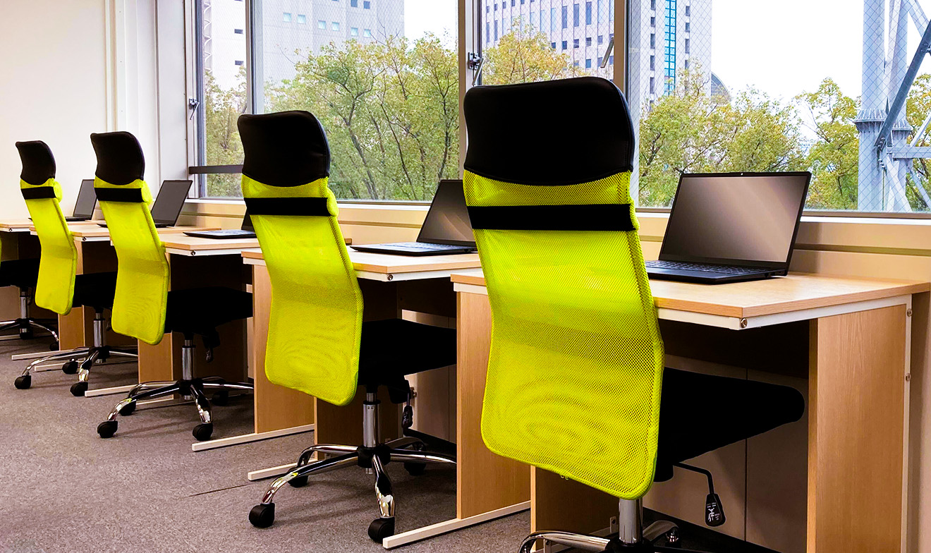 高層ビルが見える大きな窓に向かって配置された複数の机・ノートPC・黄色い椅子