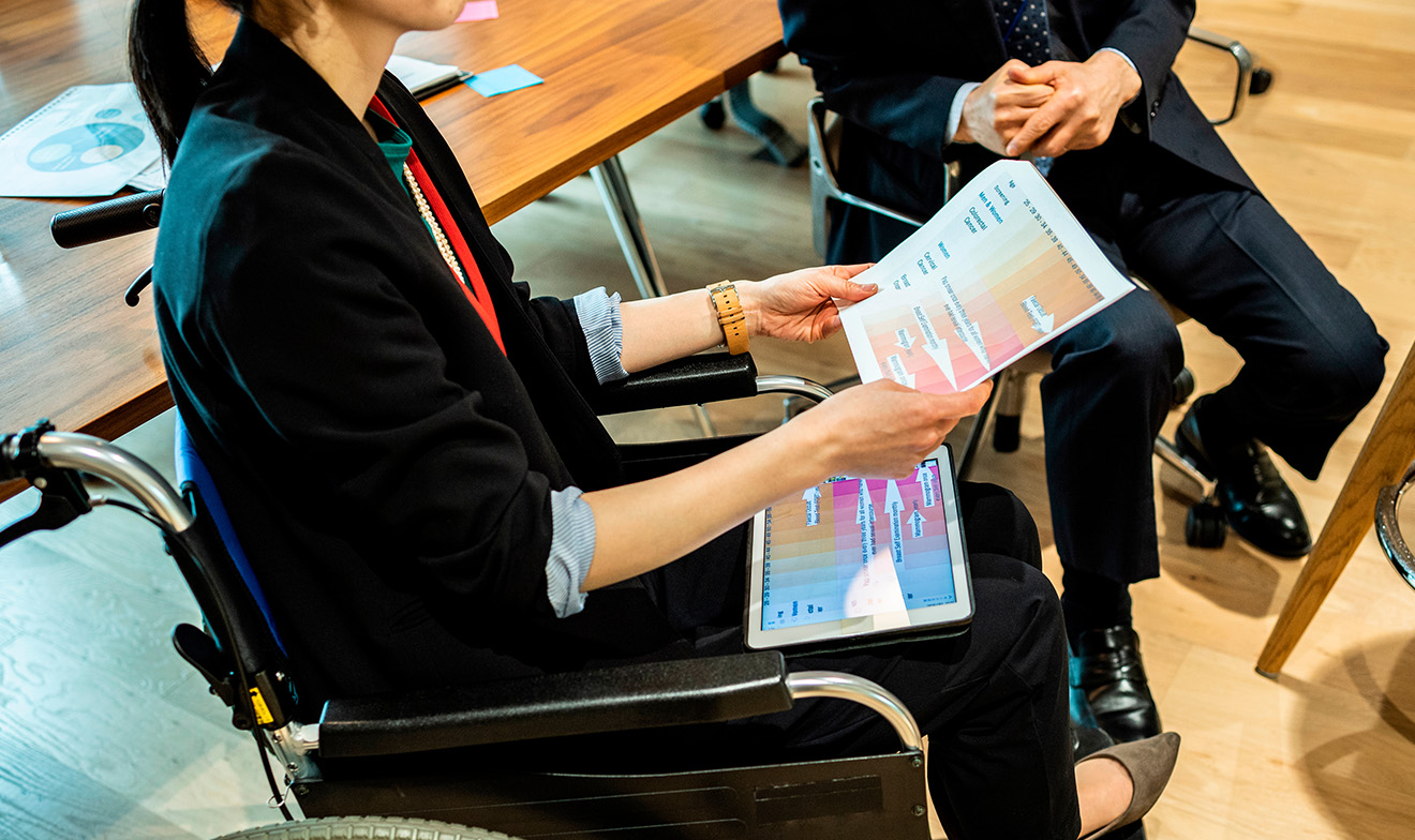 車椅子に座りグラフが印刷された紙面を両手で持ち説明する人